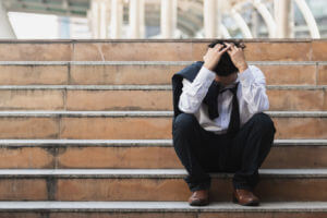 A stressed young Asian businessman in a suit sits on stairs with his hands on his head.